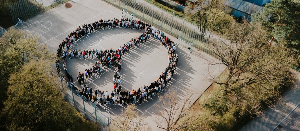 Luftaufnahme: Schülerinnen, Schüler und Lehrkräfte bilden auf dem Schulhof ein großes Friedenszeichen